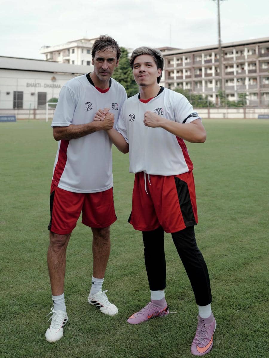 Dua pria mengenakan seragam sepak bola berwarna putih dan merah berpose di lapangan hijau dengan latar gedung bertingkat.