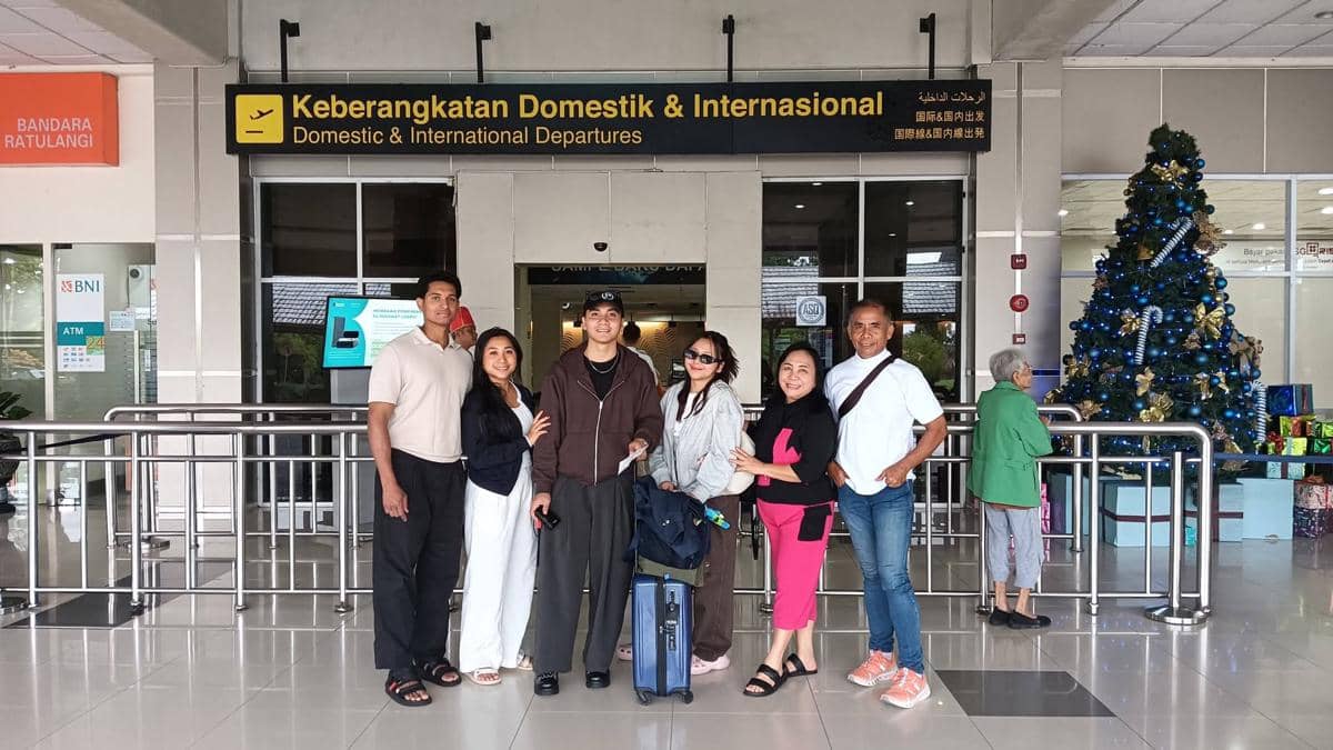 Enam orang berdiri bersama di depan area keberangkatan Bandara Internasional Sam Ratulangi Manado dengan pohon Natal di sisi kanan.