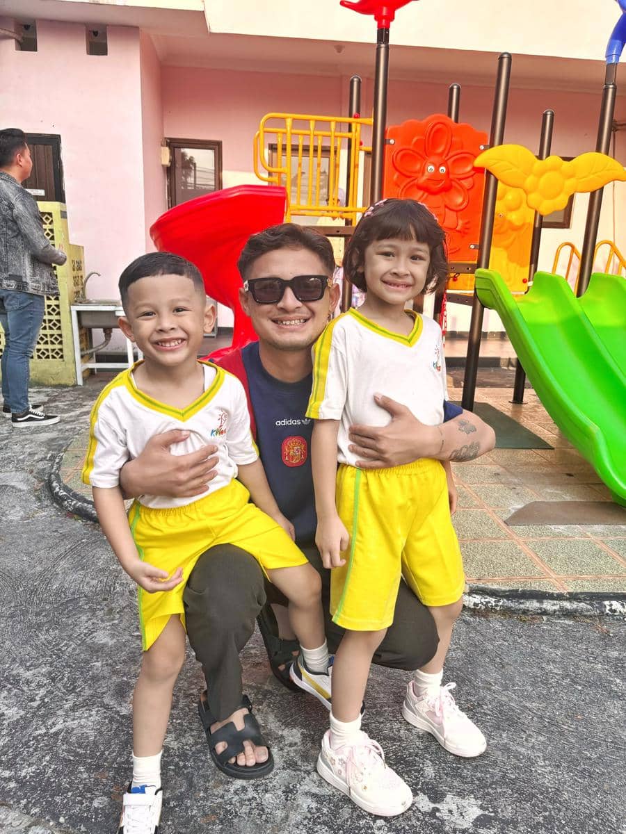 Seorang pria berpose bersama dua anak berseragam olahraga kuning di area bermain sekolah dengan perosotan warna-warni di belakang mereka.