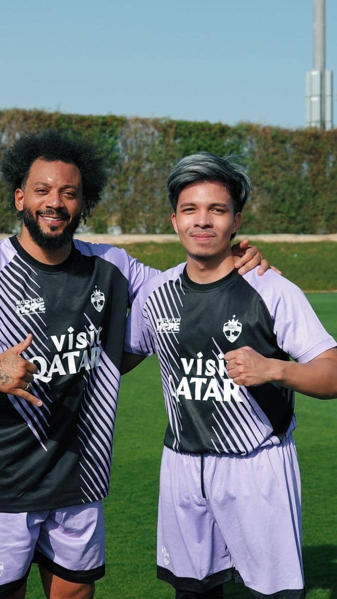 Dua pria mengenakan seragam sepak bola berwarna ungu dan hitam dengan tulisan Visit Qatar berpose bersama di lapangan hijau.