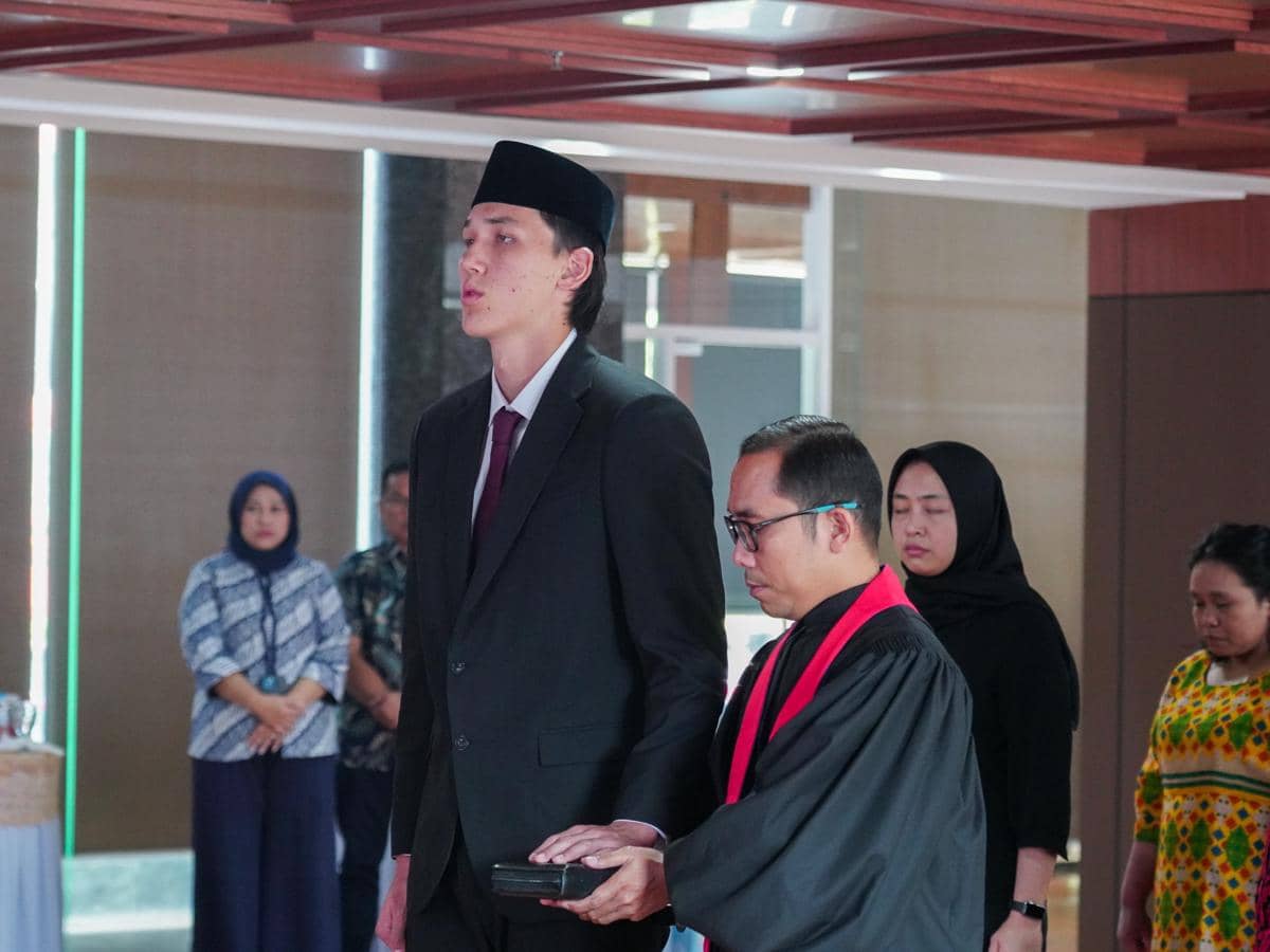 Seorang pria mengenakan setelan jas hitam dan peci sedang mengikuti prosesi resmi bersama seorang pejabat berseragam hitam di dalam ruangan.