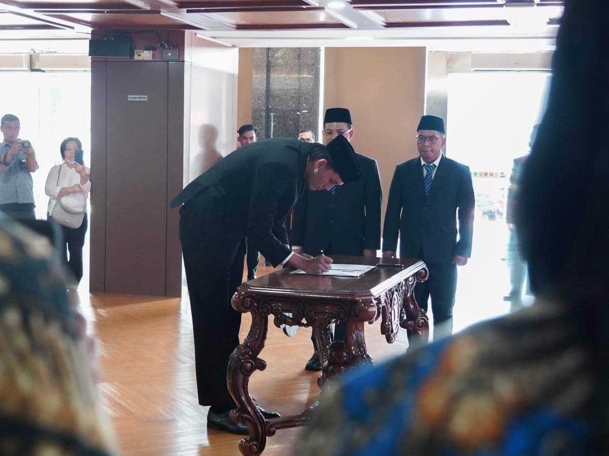 Seorang pria mengenakan setelan jas hitam dan peci menandatangani dokumen di meja kayu ukir, disaksikan beberapa orang di ruangan resmi.
