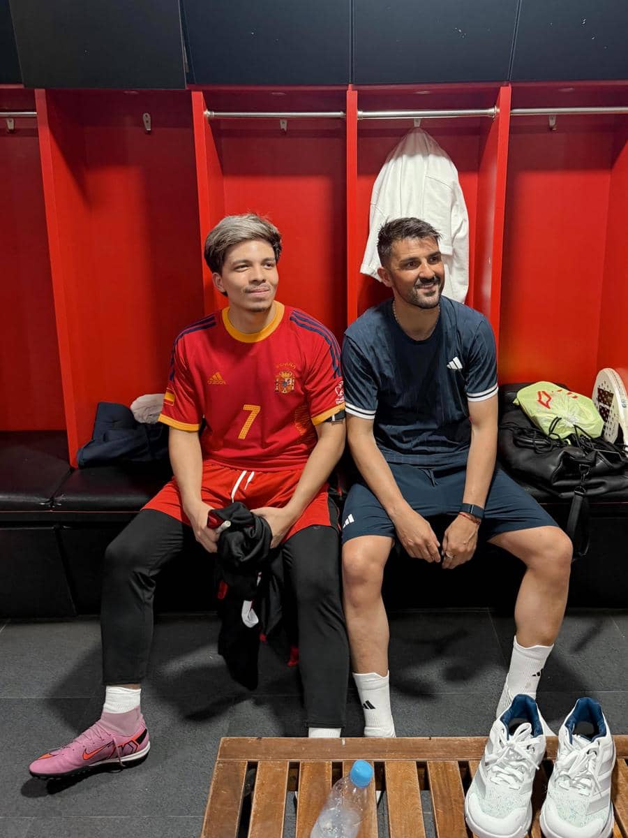 Dua pria duduk di ruang ganti berwarna merah, salah satunya mengenakan jersey sepak bola Spanyol dan yang lain mengenakan pakaian olahraga biru tua.