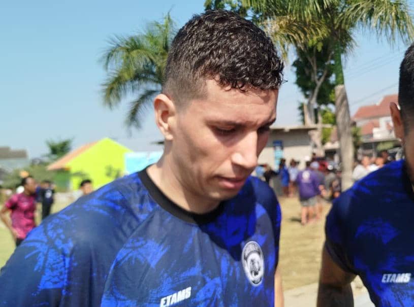Seorang pemain Arema FC mengenakan jersey biru sedang berada di area luar ruangan dengan latar pepohonan dan beberapa orang di belakangnya.