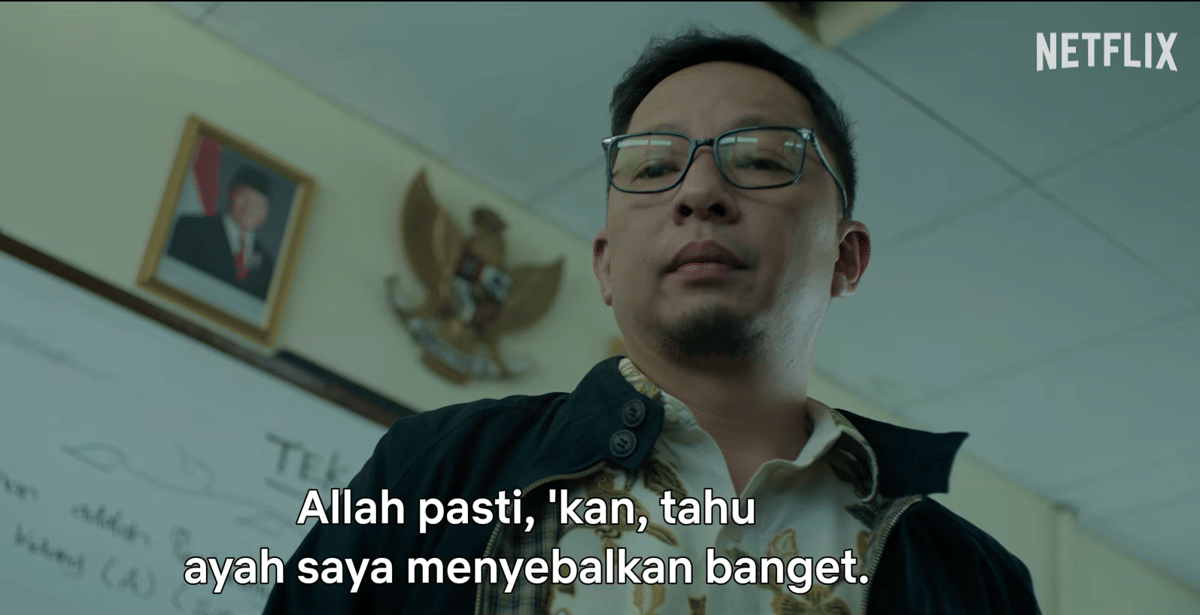 Seorang pria berkacamata berdiri di dalam ruangan dengan lambang Garuda Pancasila dan foto presiden di dinding belakangnya.