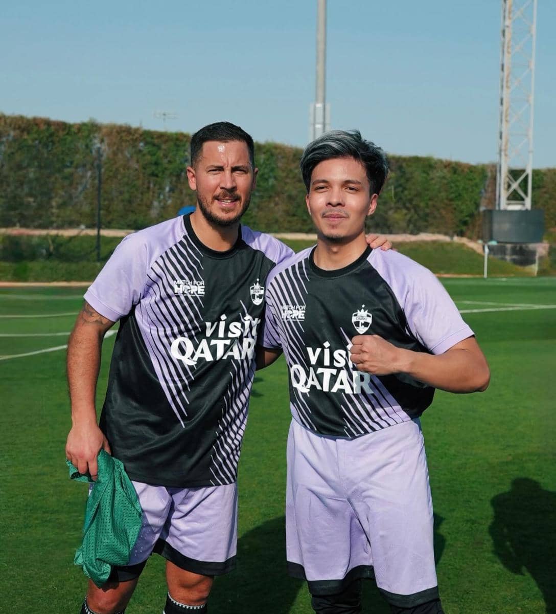 Dua pria mengenakan seragam sepak bola berwarna ungu dan hitam berpose bersama di lapangan hijau di bawah langit cerah.