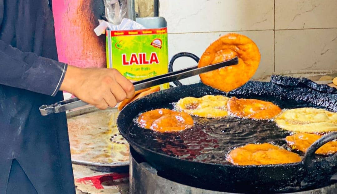 Seseorang sedang menggoreng roti bundar berwarna keemasan di wajan besar berisi minyak panas menggunakan penjepit logam.