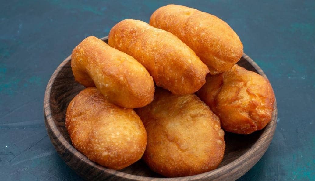 Beberapa potong roti goreng berwarna keemasan tersusun di dalam mangkuk kayu di atas permukaan meja berwarna biru gelap.