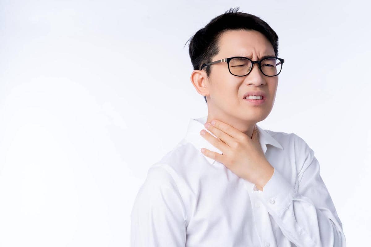 Pria mengenakan kemeja putih memegang lehernya sambil meringis kesakitan, menggambarkan rasa sakit pada tenggorokan.