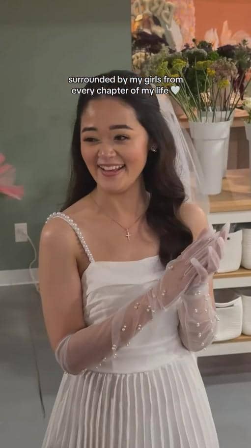 Seorang wanita mengenakan gaun putih dan sarung tangan transparan berhias mutiara tersenyum di acara bridal shower dengan latar bunga segar.
