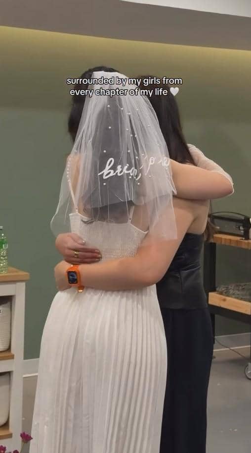 Seorang wanita mengenakan gaun putih dan veil bertuliskan 'bride' sedang berpelukan dengan temannya di acara bridal shower.