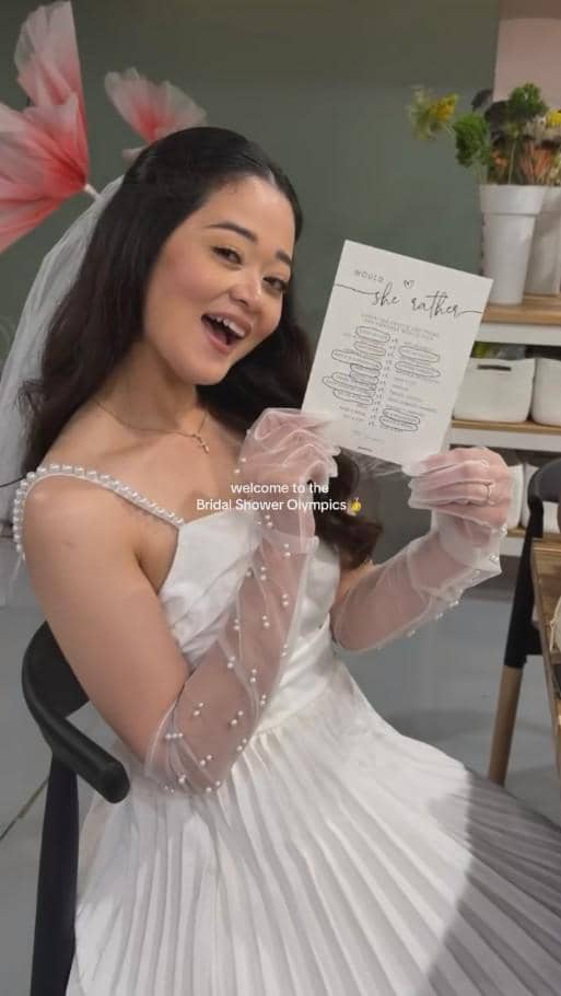 Seorang wanita mengenakan gaun putih dan sarung tangan transparan berhias mutiara tersenyum sambil memegang kartu di acara bridal shower.