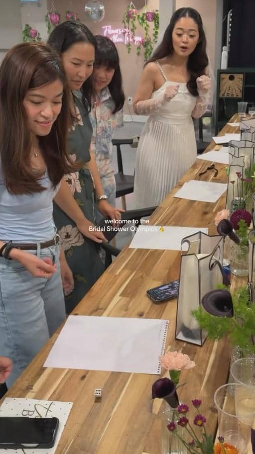 Beberapa wanita tersenyum dan bermain permainan di meja kayu panjang saat acara bridal shower dengan dekorasi bunga dan hadiah.