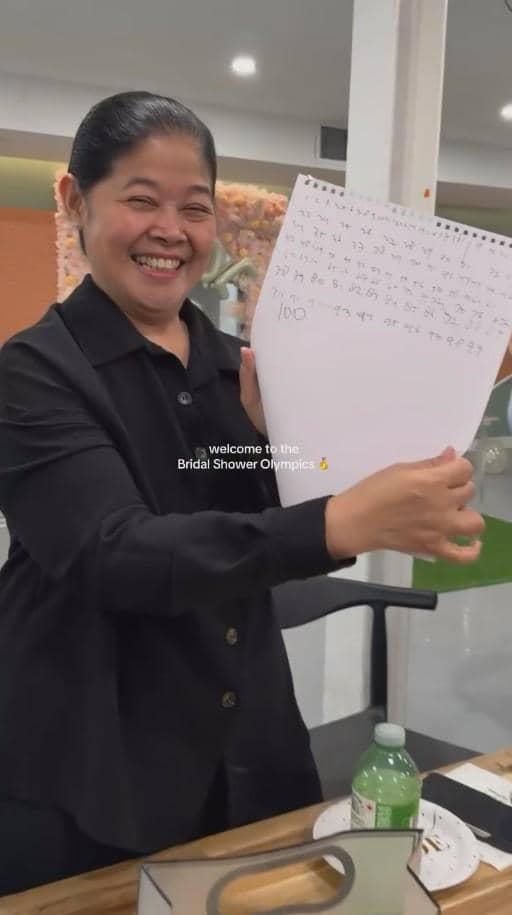 Seorang wanita tersenyum sambil memegang kertas bertuliskan angka dan huruf di acara Bridal Shower Olympics di sebuah ruangan.