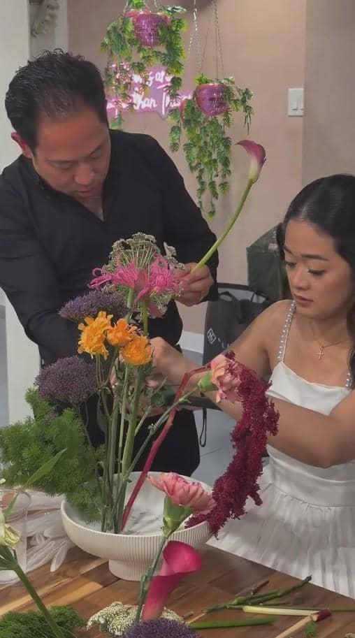 Seorang pria dan wanita sedang menyusun rangkaian bunga berwarna-warni di atas meja kayu dalam acara bridal shower.