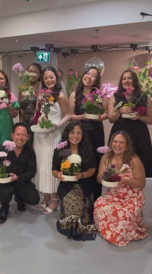 Sekelompok orang tersenyum sambil memegang rangkaian bunga dalam pot kecil pada acara bridal shower di dalam ruangan yang dihiasi bunga.