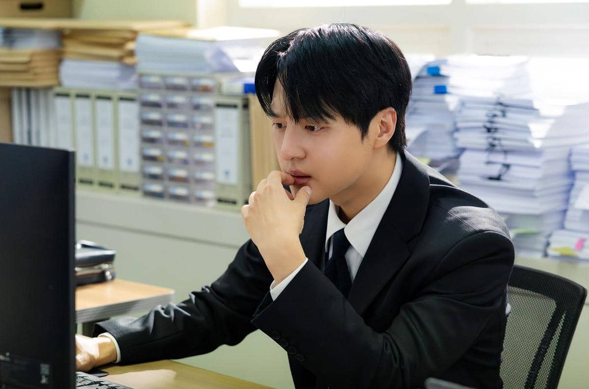 Seorang pria mengenakan setelan jas hitam sedang duduk di depan komputer di kantor dengan tumpukan berkas di belakangnya.