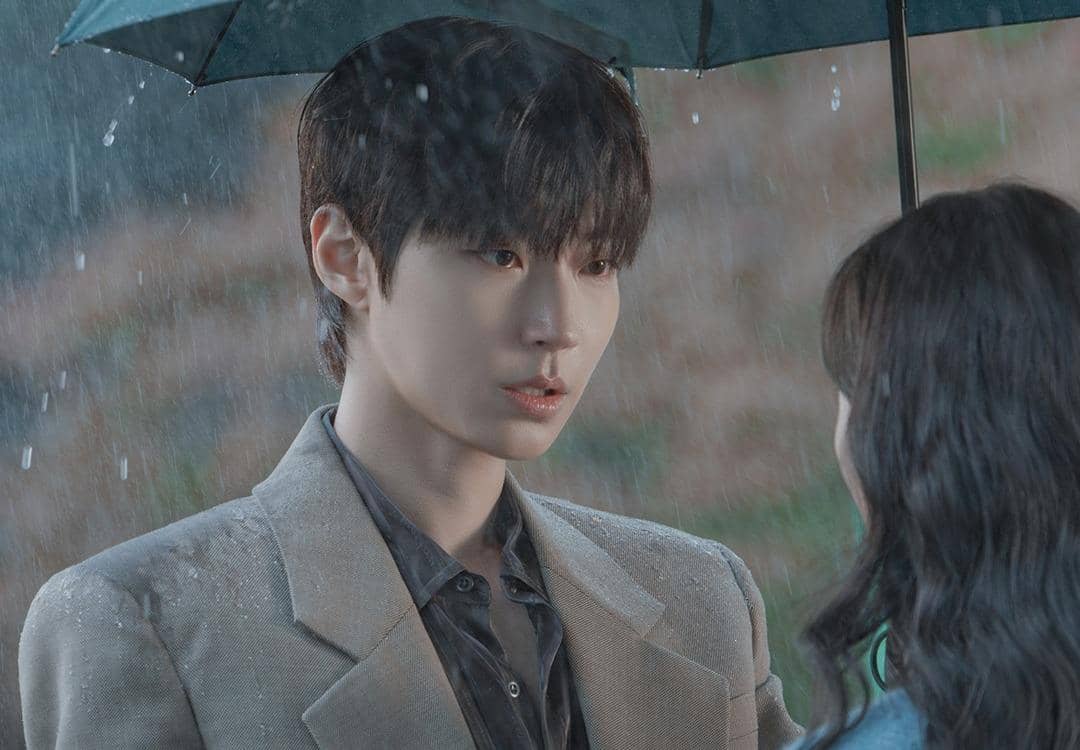 Seorang pria berjas abu-abu berdiri di bawah payung bersama seorang wanita saat hujan dalam adegan drama Korea Dream to You.