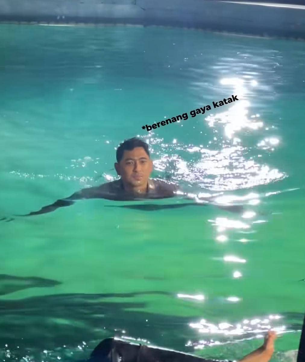 Seorang pria sedang berenang di kolam dengan air berwarna hijau kebiruan, tampak melakukan gaya katak di bawah pencahayaan terang.