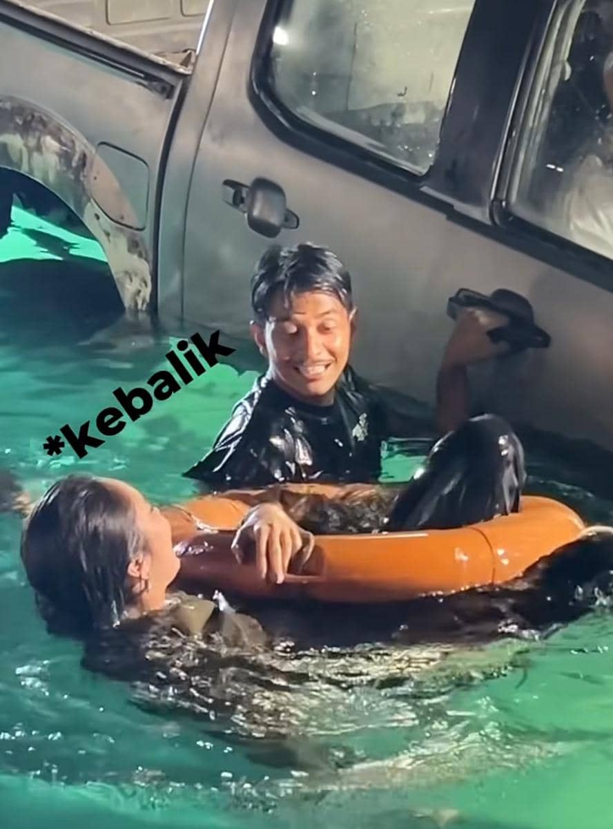Dua orang aktor berada di air dekat mobil yang sebagian terendam, salah satunya memegang pelampung oranye sambil tersenyum.
