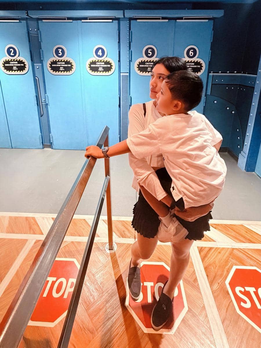 Seorang wanita mengenakan pakaian putih memeluk anak laki-lakinya di area antrean dengan pintu biru bernomor di Universal Studio Singapura.