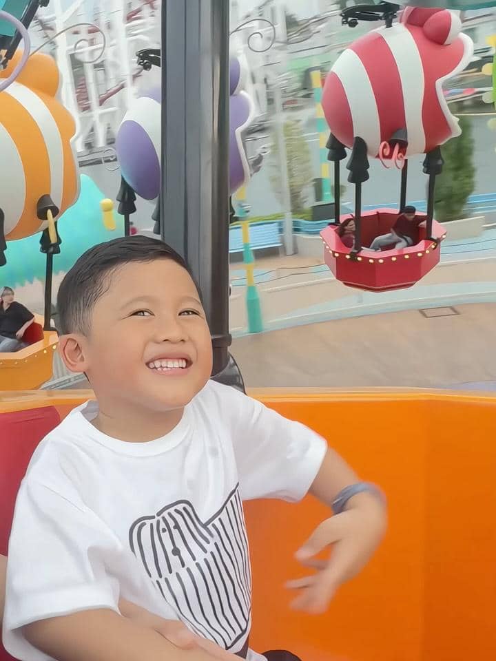 Seorang anak tersenyum ceria saat menaiki wahana berwarna cerah di Universal Studio Singapura dengan latar balon udara mainan.