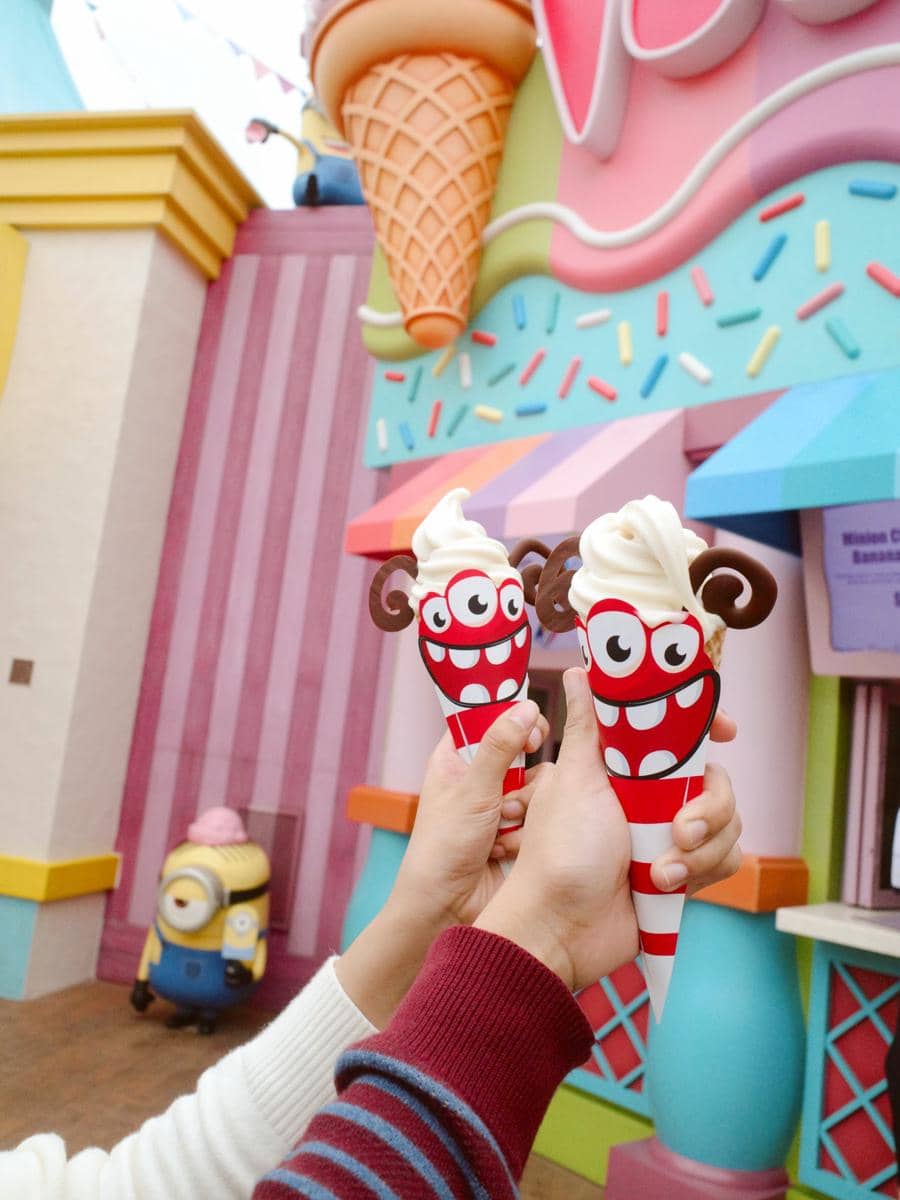 Dua tangan memegang es krim berbentuk lucu dengan cone bergambar karakter tersenyum di area warna-warni Universal Studio Singapura.