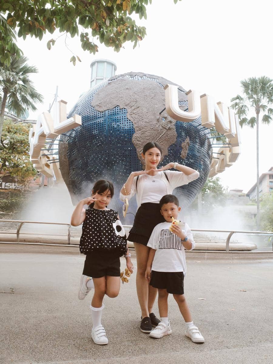 Seorang wanita berpose bersama dua anak kecil di depan bola dunia ikonik Universal Studios Singapura dengan suasana cerah dan santai.
