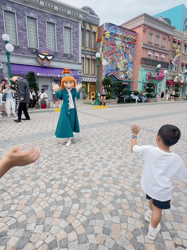Seorang anak kecil berinteraksi dengan karakter animasi di area jalan berbatu Universal Studios Singapura dengan latar bangunan warna-warni.