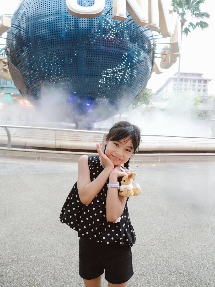 Seorang anak perempuan berpose ceria sambil memegang boneka kecil di depan bola dunia ikonik Universal Studios Singapura.