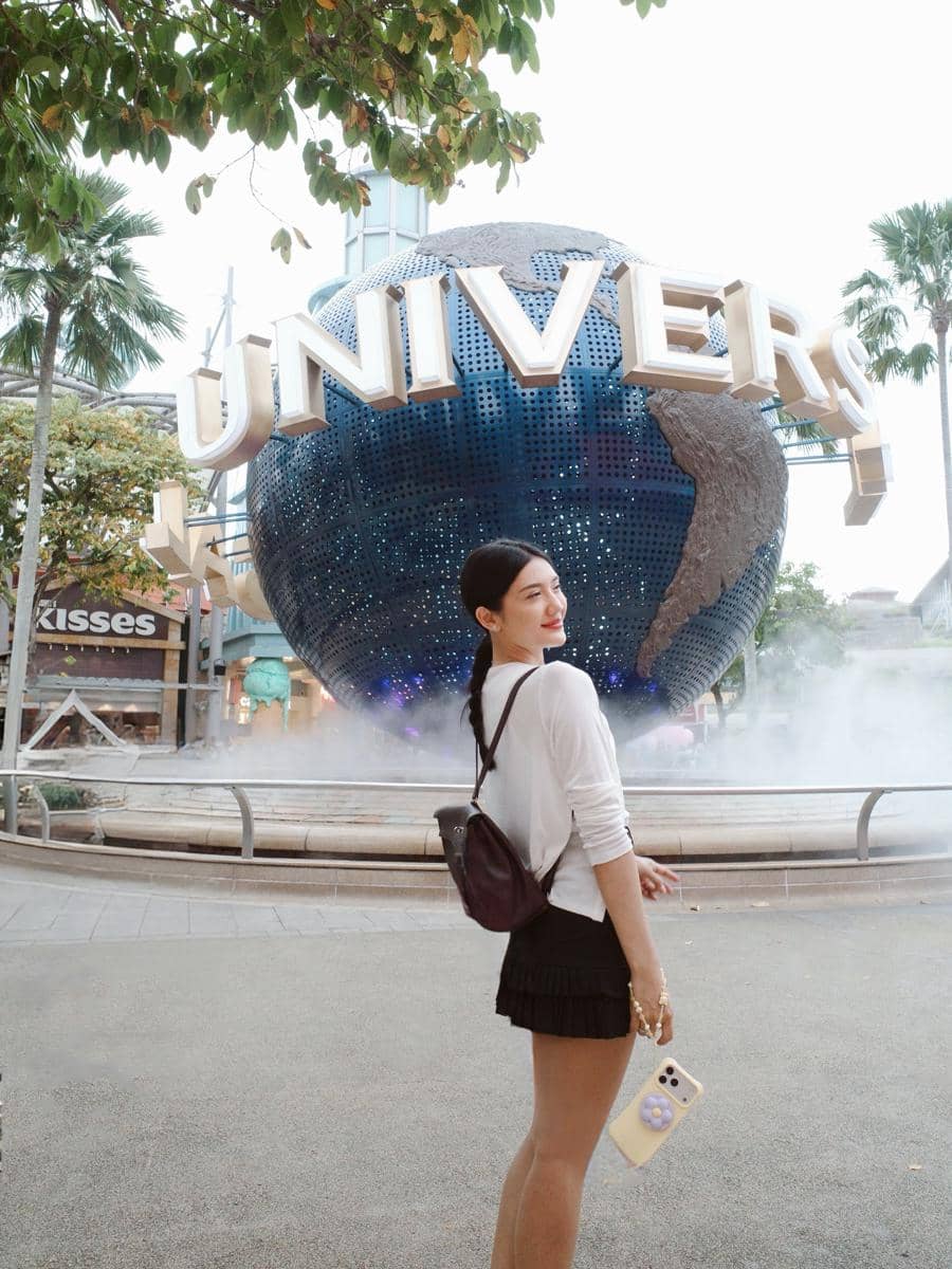 Seorang wanita berpose di depan bola dunia ikonik Universal Studios Singapura dengan latar pepohonan dan suasana cerah.
