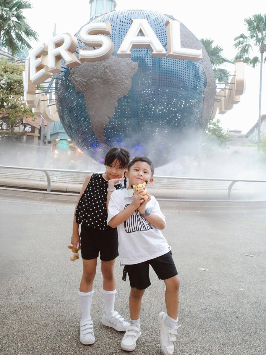 Dua anak kecil berpose ceria di depan bola dunia ikonik Universal Studios Singapura dengan suasana kabut tipis di sekitarnya.