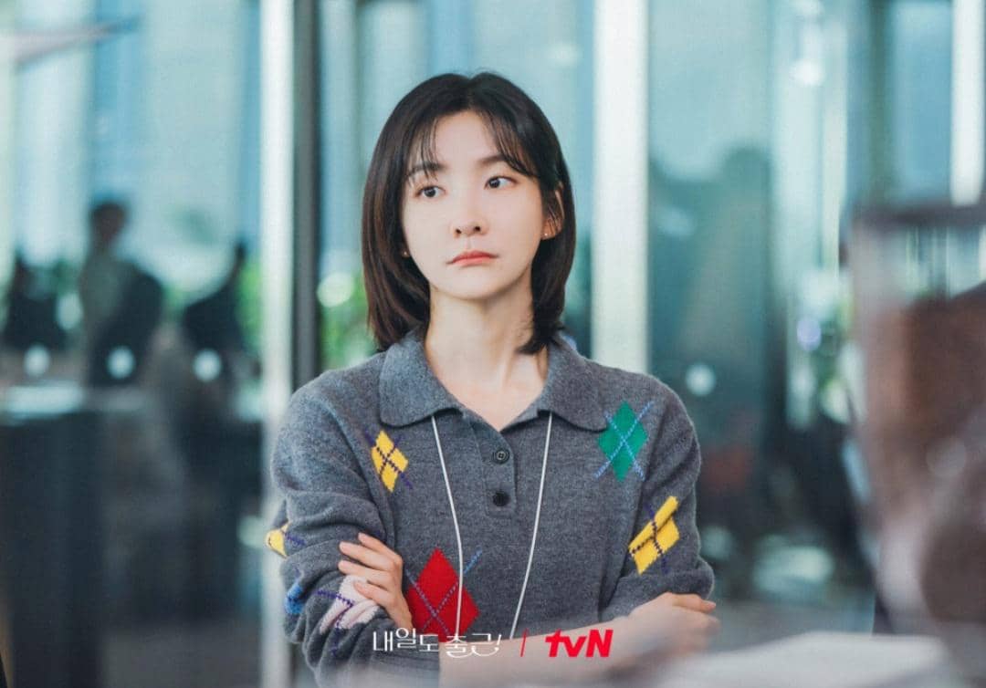 Seorang wanita berambut pendek mengenakan sweter abu-abu bermotif argyle berdiri dengan tangan terlipat di lingkungan kantor modern.