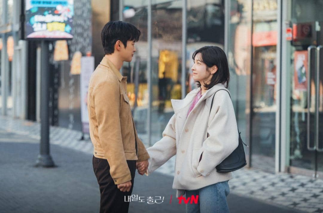 Dua karakter dalam drama Korea See You at Work Tomorrow berdiri saling menatap dan berpegangan tangan di jalan kota.