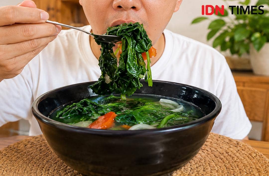 Seseorang mengenakan kaus putih sedang menyantap sayur bayam panas dari mangkuk hitam besar berisi bayam, wortel, dan kuah bening.