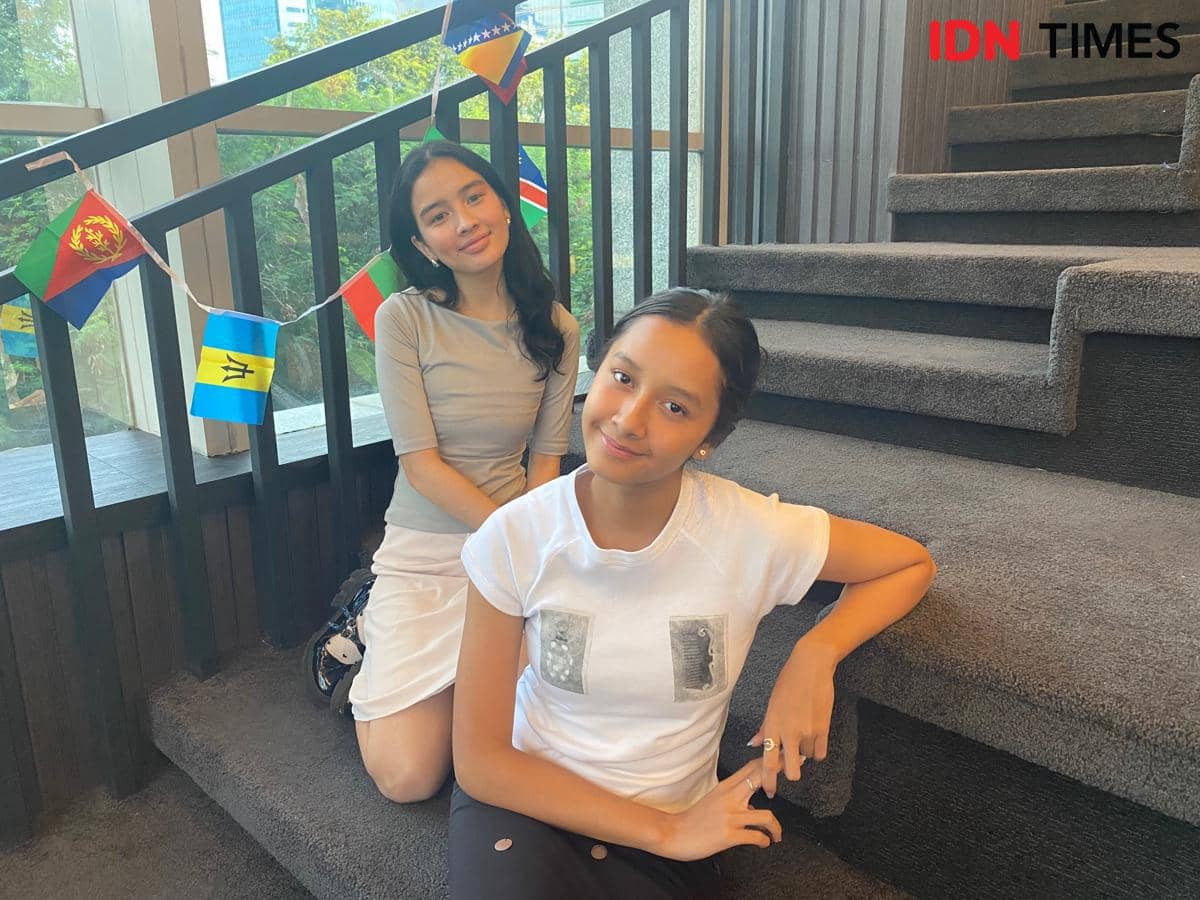 Dua perempuan berpose santai di tangga kantor dengan dekorasi bendera kecil berwarna-warni di latar belakang, suasana cerah di Jakarta.