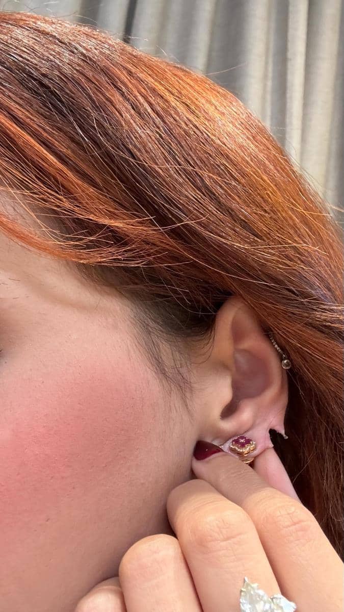 Tampak dekat telinga seorang perempuan berambut cokelat kemerahan dengan anting berhiaskan batu merah muda dan cincin besar di jarinya.