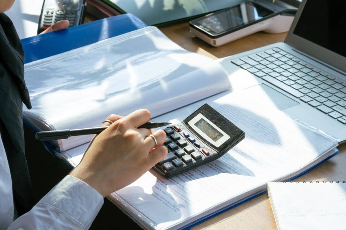 Seseorang menghitung data keuangan menggunakan kalkulator di meja kerja dengan laptop, dokumen, dan buku catatan terbuka.