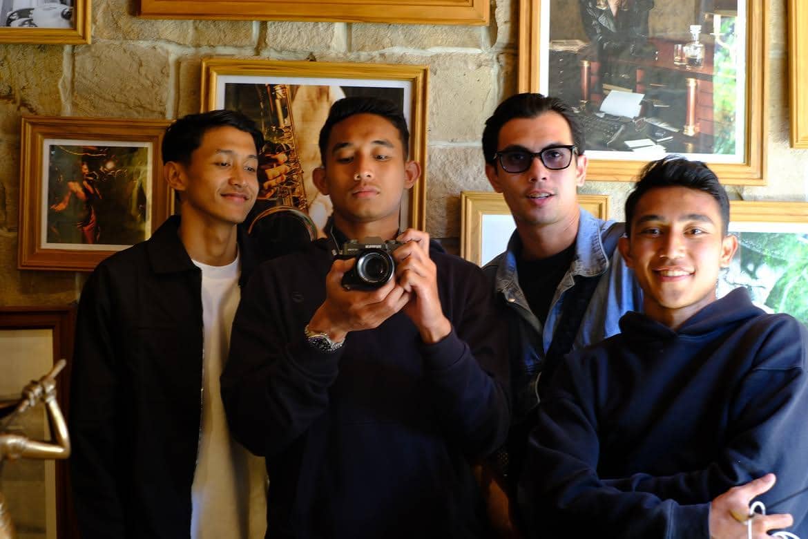 Empat pria berpose bersama di depan dinding dengan bingkai foto, salah satunya memegang kamera saat berwisata di Bali.