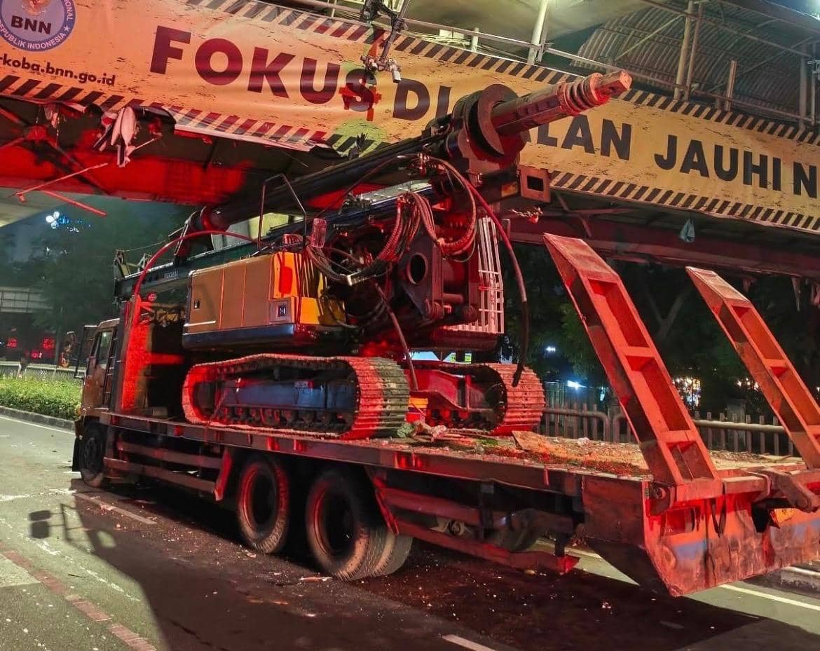 Truk pengangkut crane mengalami kecelakaan di bawah jembatan penyeberangan dengan bagian alat berat tersangkut pada struktur atas.