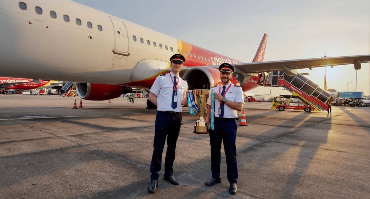 Dua pilot Vietjet berdiri di depan pesawat maskapai di landasan dengan memegang trofi besar pada sore hari.