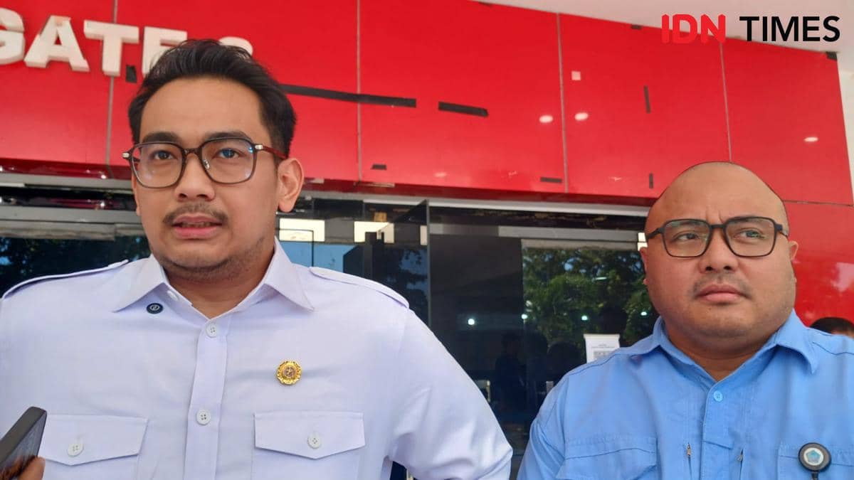 Dua pria mengenakan kemeja formal berdiri di depan gedung berwarna merah dengan ekspresi serius, salah satunya memegang ponsel.