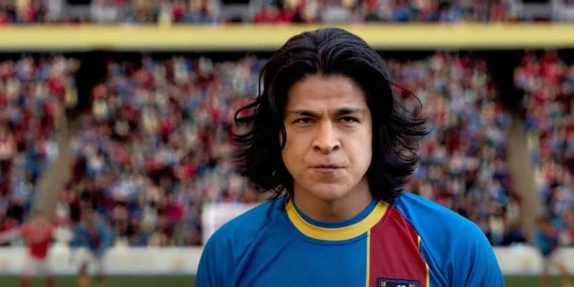 Seorang pemain sepak bola berambut panjang mengenakan seragam biru dan merah berdiri di lapangan dengan latar tribun penonton penuh warna.