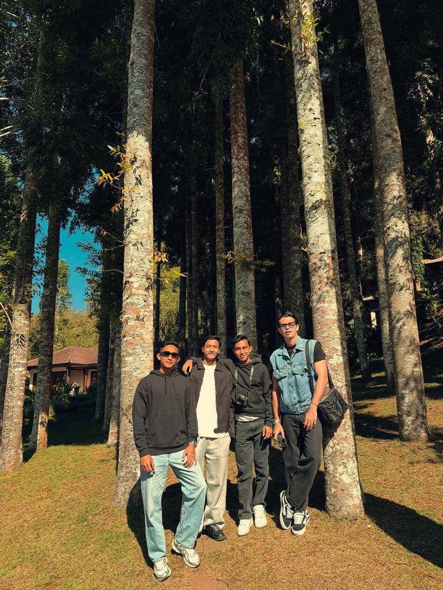 Empat pria berdiri bersama di antara pepohonan tinggi di area hutan dengan suasana cerah dan latar belakang rumah kayu di Bali.