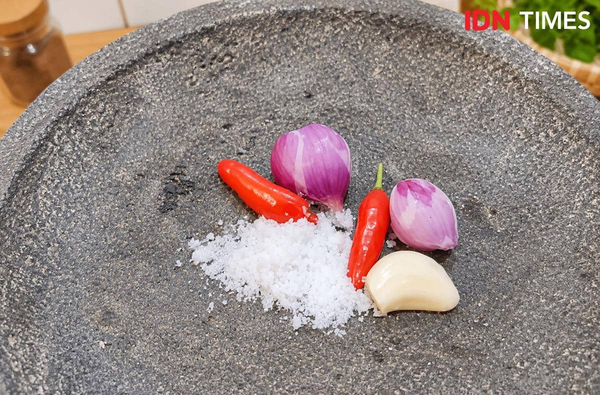 Bawang merah, cabai merah, bawang putih, dan garam kasar tersusun di atas cobek batu sebagai bumbu dasar sayur bayem geprek.