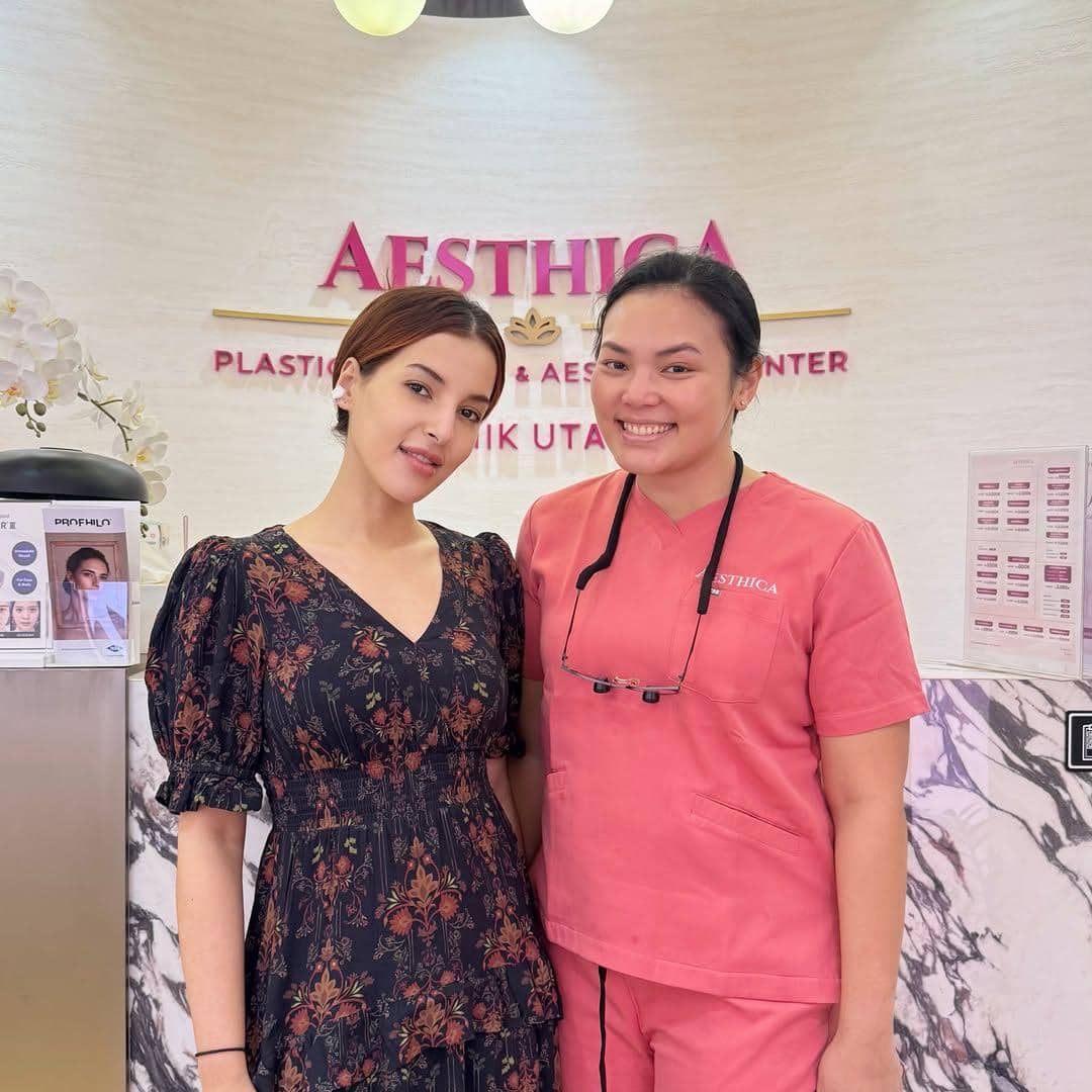 Seorang wanita bergaun hitam bermotif bunga berfoto bersama tenaga medis berseragam merah muda di klinik kecantikan Aesthica.