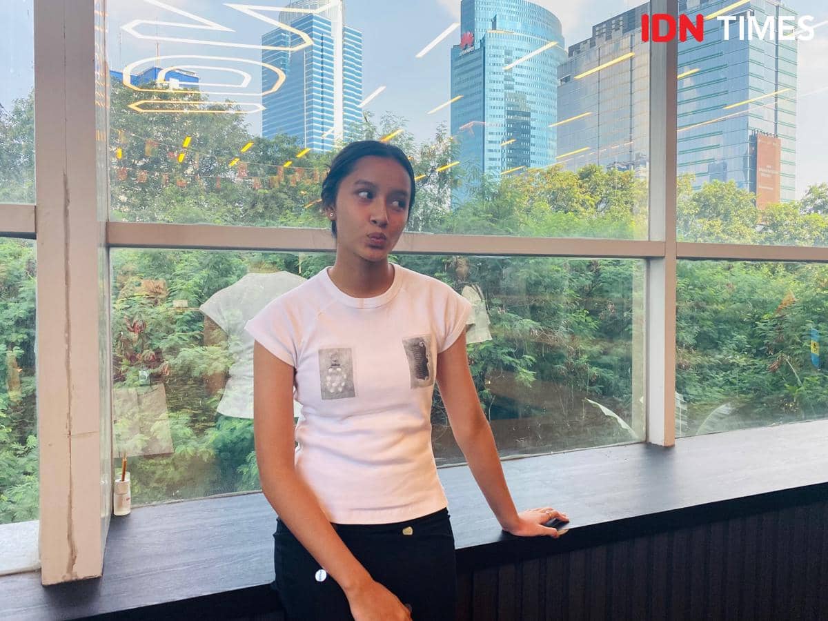 Seorang perempuan berdiri santai di dekat jendela besar dengan latar gedung tinggi dan pepohonan di kawasan perkantoran Jakarta.