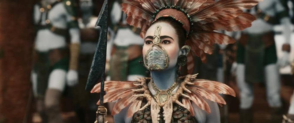 Seorang karakter perempuan berkulit biru mengenakan hiasan kepala berbulu dan topeng logam, memegang tombak dalam cuplikan film Avengers: Doomsday.