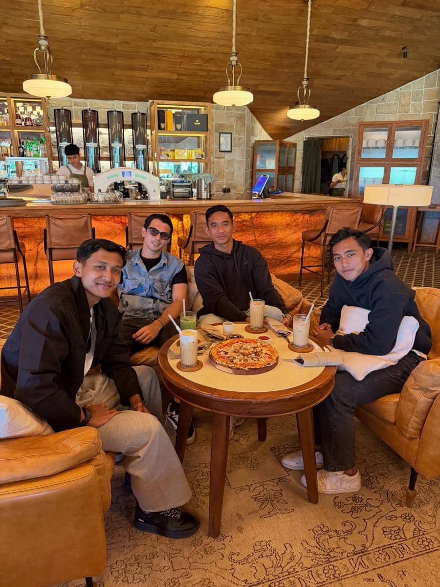 Empat pria duduk santai di kafe bergaya rustic di Bali, menikmati pizza dan minuman sambil tersenyum ke arah kamera.