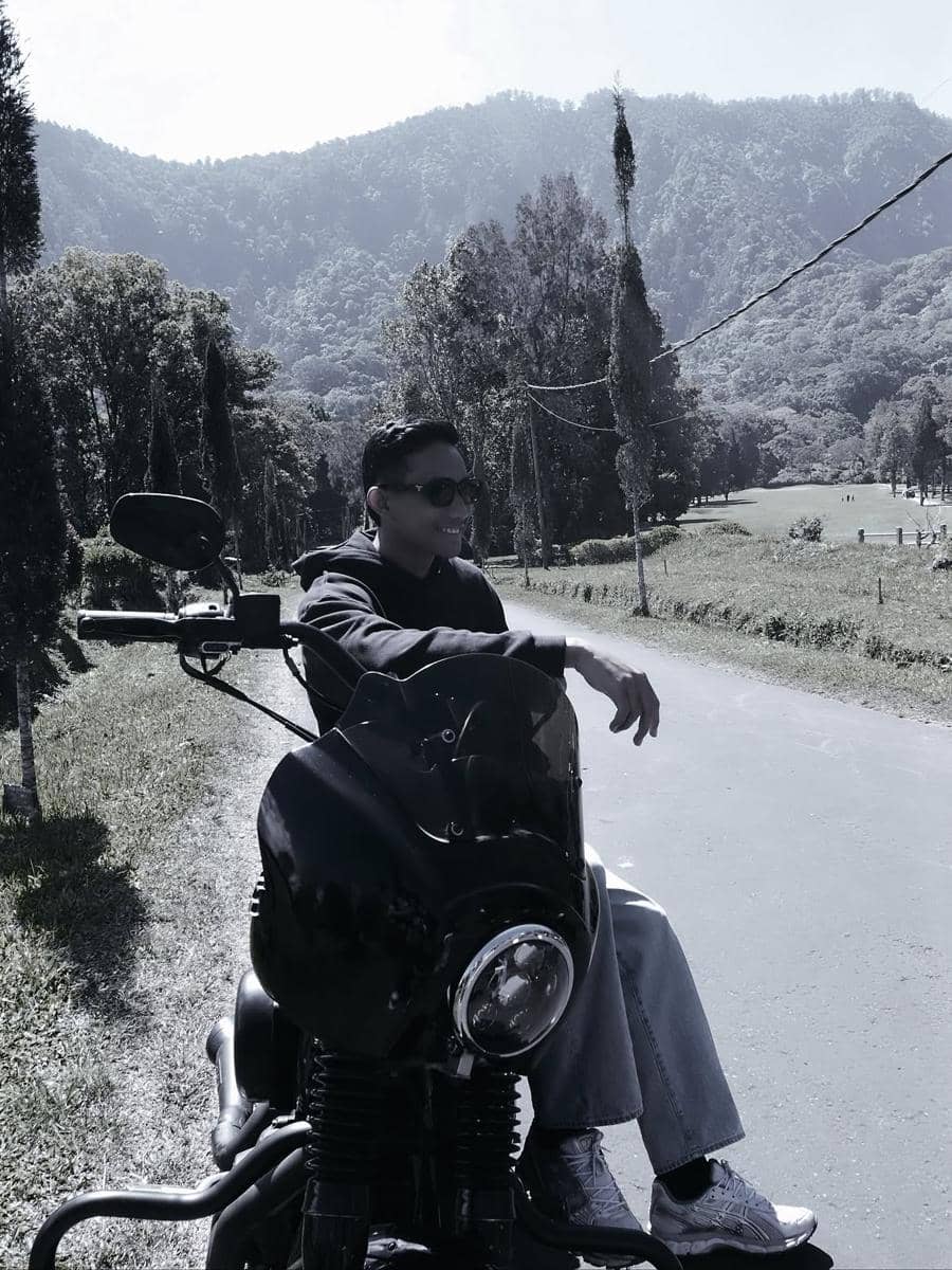Seorang pria duduk santai di atas motor besar di jalan pedesaan dengan latar pegunungan hijau di Bali pada siang hari.